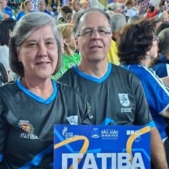Casal representante de Itatiba posa sorridente em meio à torcida durante os Jogos da Melhor Idade, segurando uma placa com o nome da cidade.