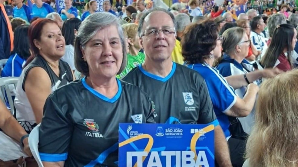 Casal representante de Itatiba posa sorridente em meio à torcida durante os Jogos da Melhor Idade, segurando uma placa com o nome da cidade.