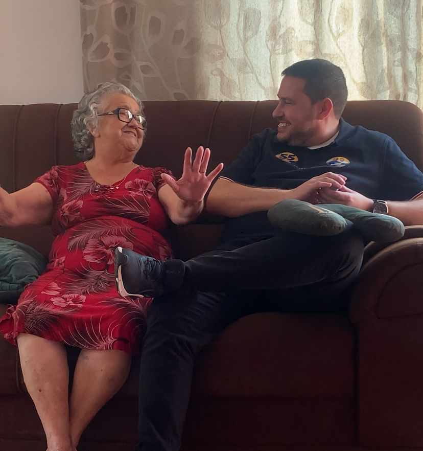 João Paulo sentado em um sofá, sorrindo e conversando com uma senhora idosa de óculos em um ambiente doméstico iluminado.