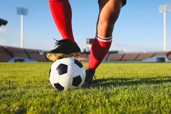 Jogador de futebol com uniforme vermelho e chuteiras pretas domina a bola em campo de gramado, durante partida em estádio iluminado.