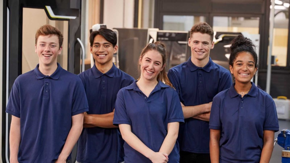 Cinco jovens aprendizes uniformizados com camisetas azul-marinho sorrindo em ambiente de trabalho, simbolizando estágio e programas de capacitação profissional. Cinco jovens aprendizes uniformizados com camisetas azul-marinho sorrindo em ambiente de trabalho, simbolizando estágio e programas de capacitação profissional.