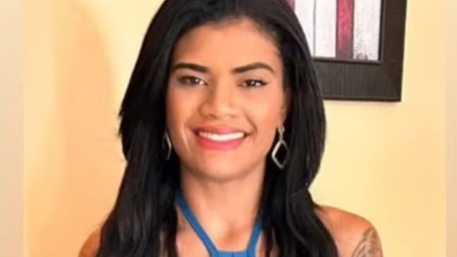 Jovem de cabelos longos e pretos, sorridente, usando brincos prateados e blusa azul, em frente a uma parede clara com quadro colorido ao fundo.