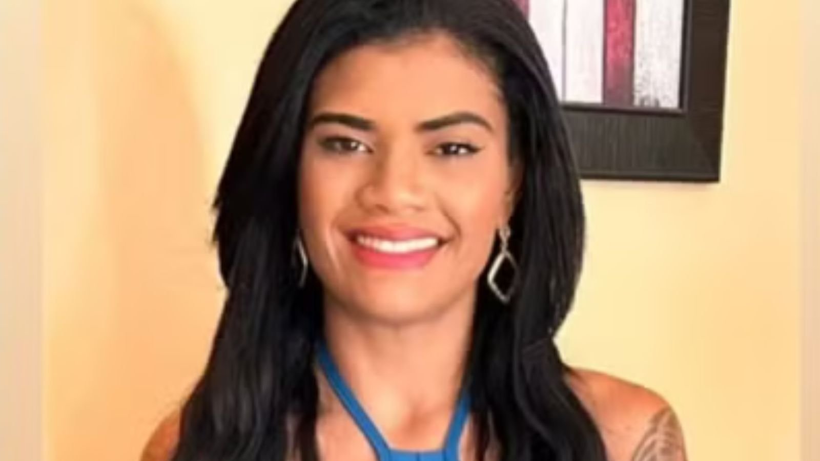 Jovem de cabelos longos e pretos, sorridente, usando brincos prateados e blusa azul, em frente a uma parede clara com quadro colorido ao fundo.