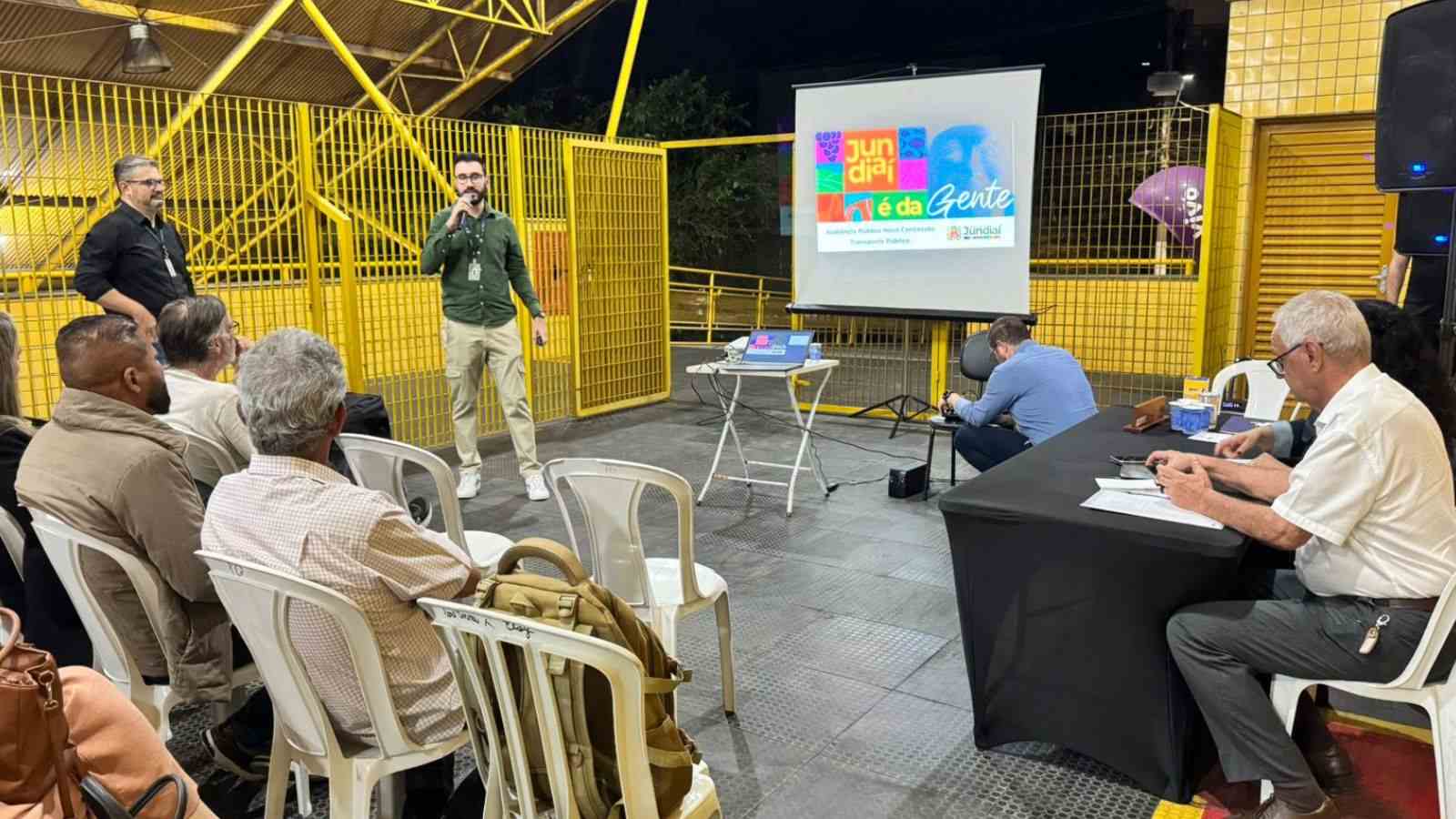 Apresentação da Secretaria de Mobilidade e Transporte durante audiência pública no Terminal Colônia, com moradores assistindo e tela projetando o logotipo “Jundiaí é da Gente