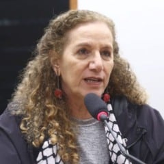 Mulher de cabelos cacheados fala ao microfone durante uma sessão em ambiente interno, vestindo casaco escuro e lenço preto e branco. Mulher de cabelos cacheados fala ao microfone durante uma sessão em ambiente interno, vestindo casaco escuro e lenço preto e branco.