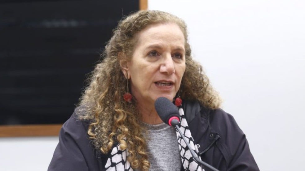 Mulher de cabelos cacheados fala ao microfone durante uma sessão em ambiente interno, vestindo casaco escuro e lenço preto e branco.