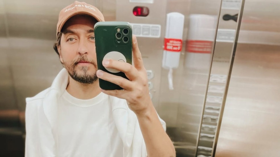 Homem de barba curta e boné bege tira uma selfie em frente ao espelho de um elevador, segurando o celular verde com suporte redondo branco. Homem de barba curta e boné bege tira uma selfie em frente ao espelho de um elevador, segurando o celular verde com suporte redondo branco.