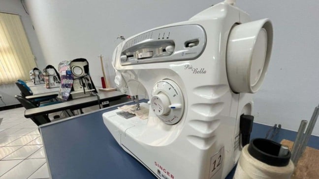 Máquina de costura Singer em sala de aula, com outras máquinas alinhadas ao fundo e um carretel de linha em destaque sobre a mesa.