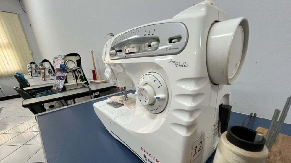 Máquina de costura Singer em sala de aula, com outras máquinas alinhadas ao fundo e um carretel de linha em destaque sobre a mesa.