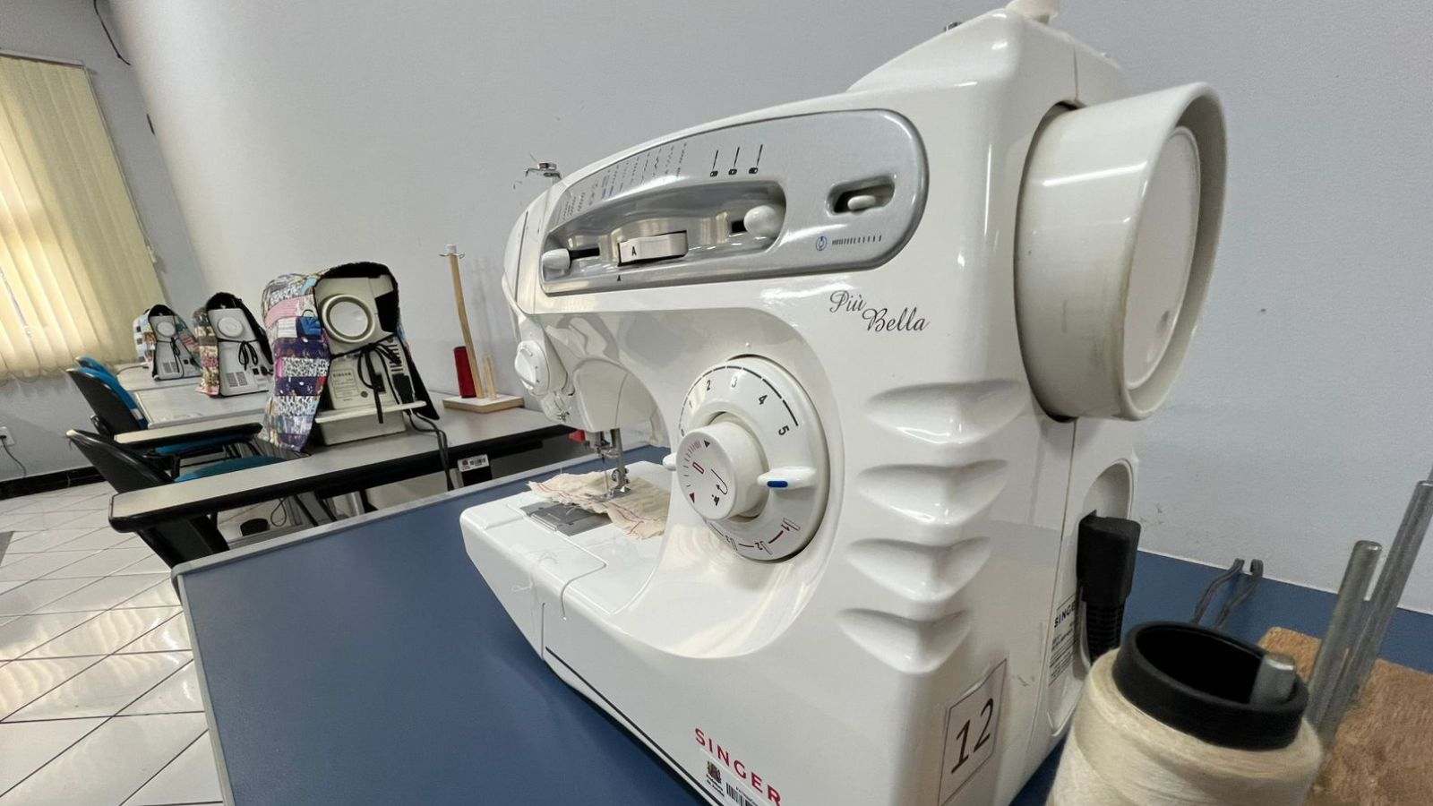 Máquina de costura Singer em sala de aula, com outras máquinas alinhadas ao fundo e um carretel de linha em destaque sobre a mesa.