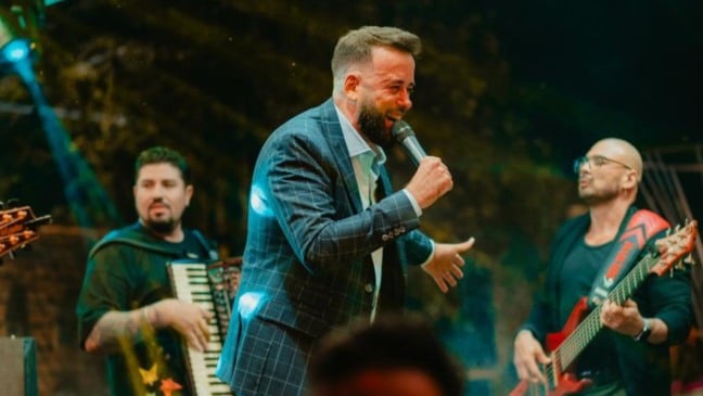 Cantor Marcelo Gut se apresenta no palco com banda ao fundo, transmitindo energia e alegria.