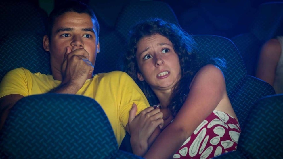 Casal assustado assistindo a um filme de terror no cinema, reagindo com medo e expressão de pavor.