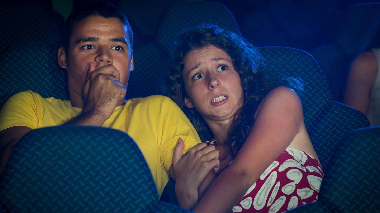 Casal assustado assistindo a um filme de terror no cinema, reagindo com medo e expressão de pavor.