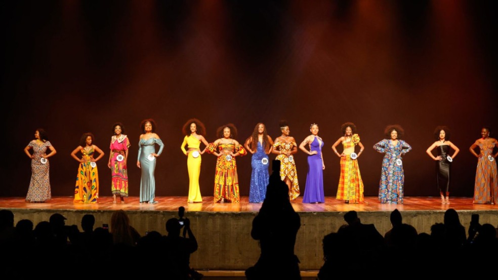Candidatas do concurso Miss Pérola Negra desfilam no palco com vestidos afro coloridos, celebrando a beleza negra.