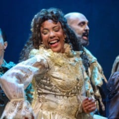 Elenco do musical “Dom Casmurro” em cena, com atores vestidos com figurinos de época e expressões alegres durante apresentação no palco.