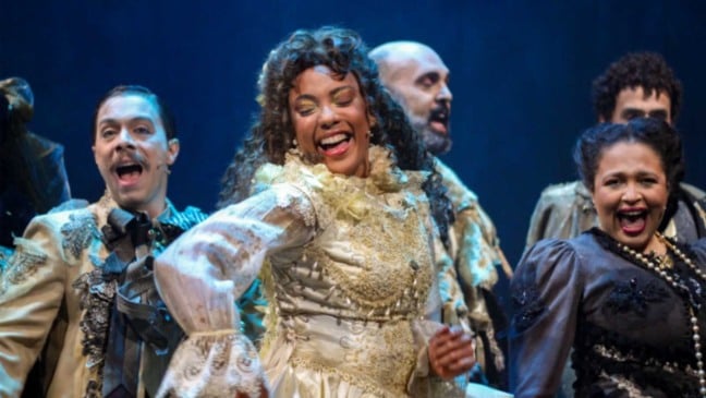 Elenco do musical “Dom Casmurro” em cena, com atores vestidos com figurinos de época e expressões alegres durante apresentação no palco. Elenco do musical “Dom Casmurro” em cena, com atores vestidos com figurinos de época e expressões alegres durante apresentação no palco.