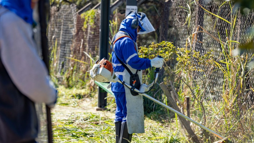 Funcionário com uniforme azul e equipamentos de proteção usa roçadeira para cortar mato em área pública durante o mutirão “Quem Ama, Cuida” em Jundiaí. Funcionário com uniforme azul e equipamentos de proteção usa roçadeira para cortar mato em área pública durante o mutirão “Quem Ama, Cuida” em Jundiaí.