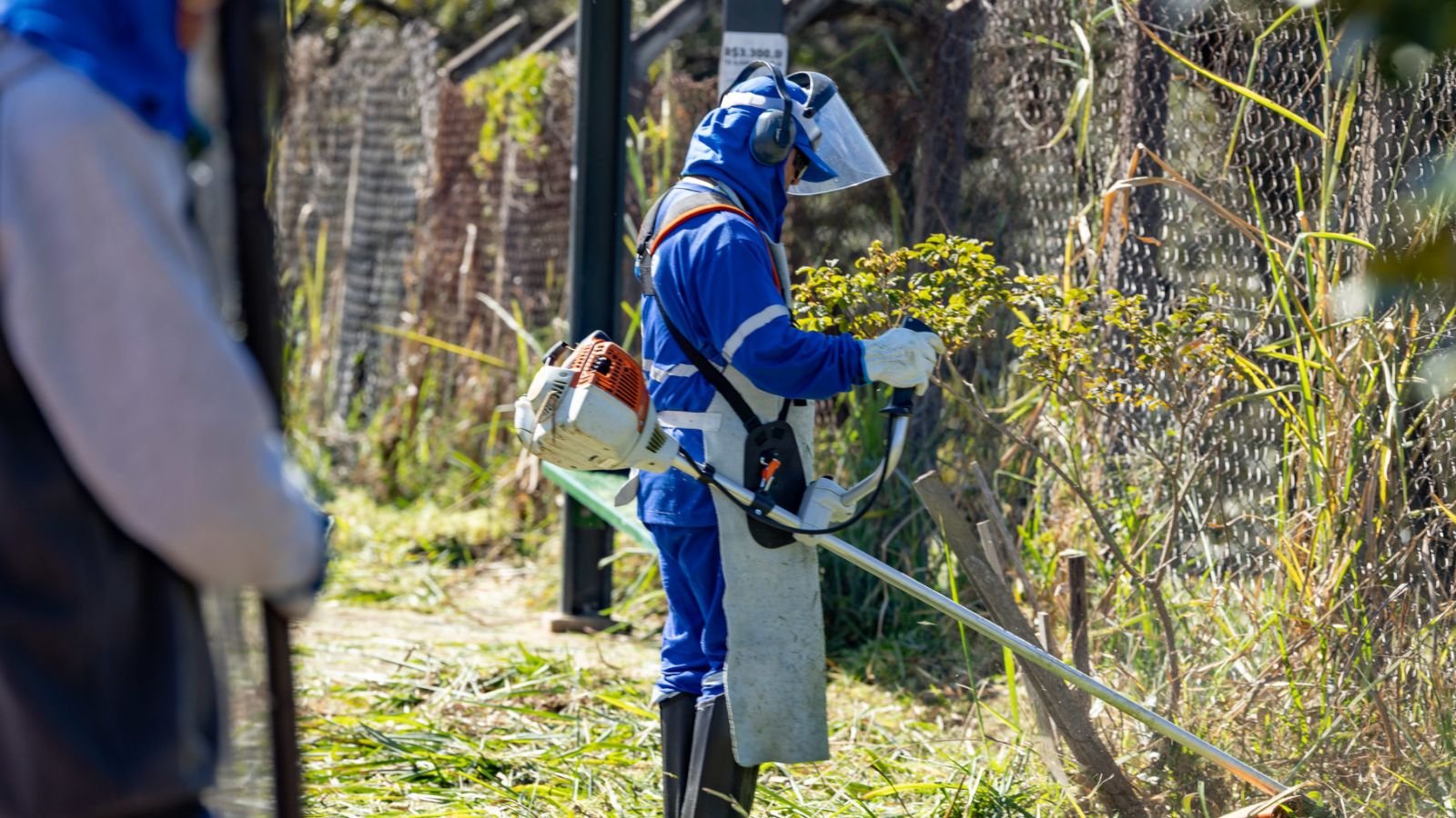 Funcionário com uniforme azul e equipamentos de proteção usa roçadeira para cortar mato em área pública durante o mutirão “Quem Ama, Cuida” em Jundiaí.