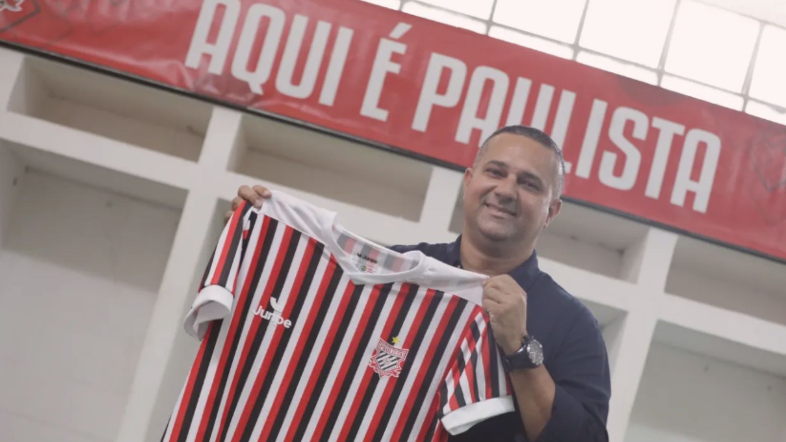 Novo técnico do Paulista FC, Raphael Pereira sorri enquanto segura a camisa tricolor do clube em sua apresentação no Estádio Dr. Jayme Cintra.