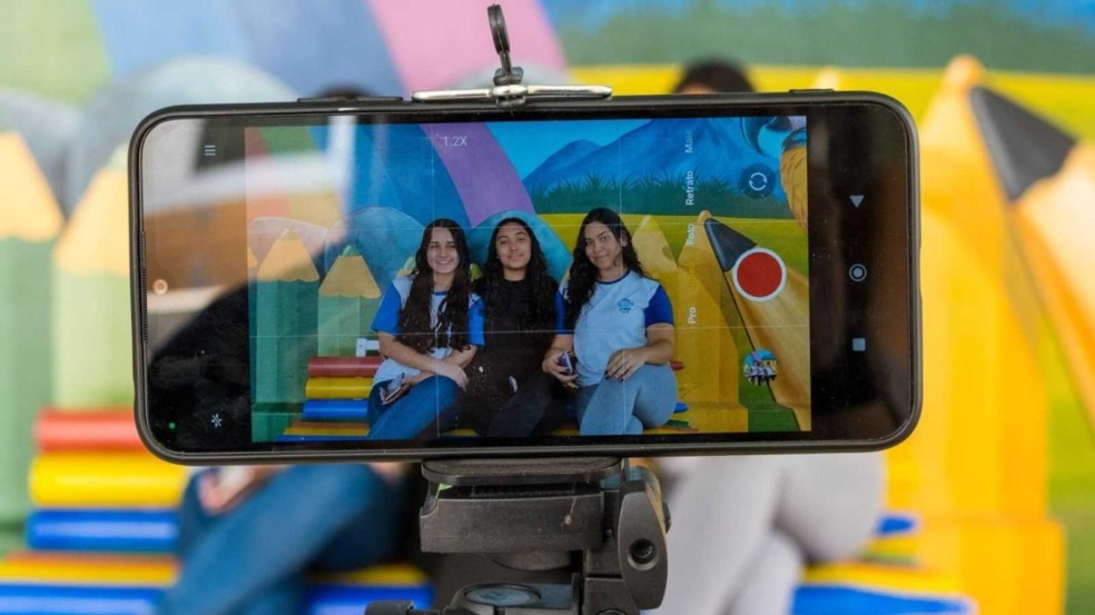 Celular em tripé grava três jovens estudantes sorrindo em frente a um mural colorido durante oficina de produção audiovisual em Jundiaí.
