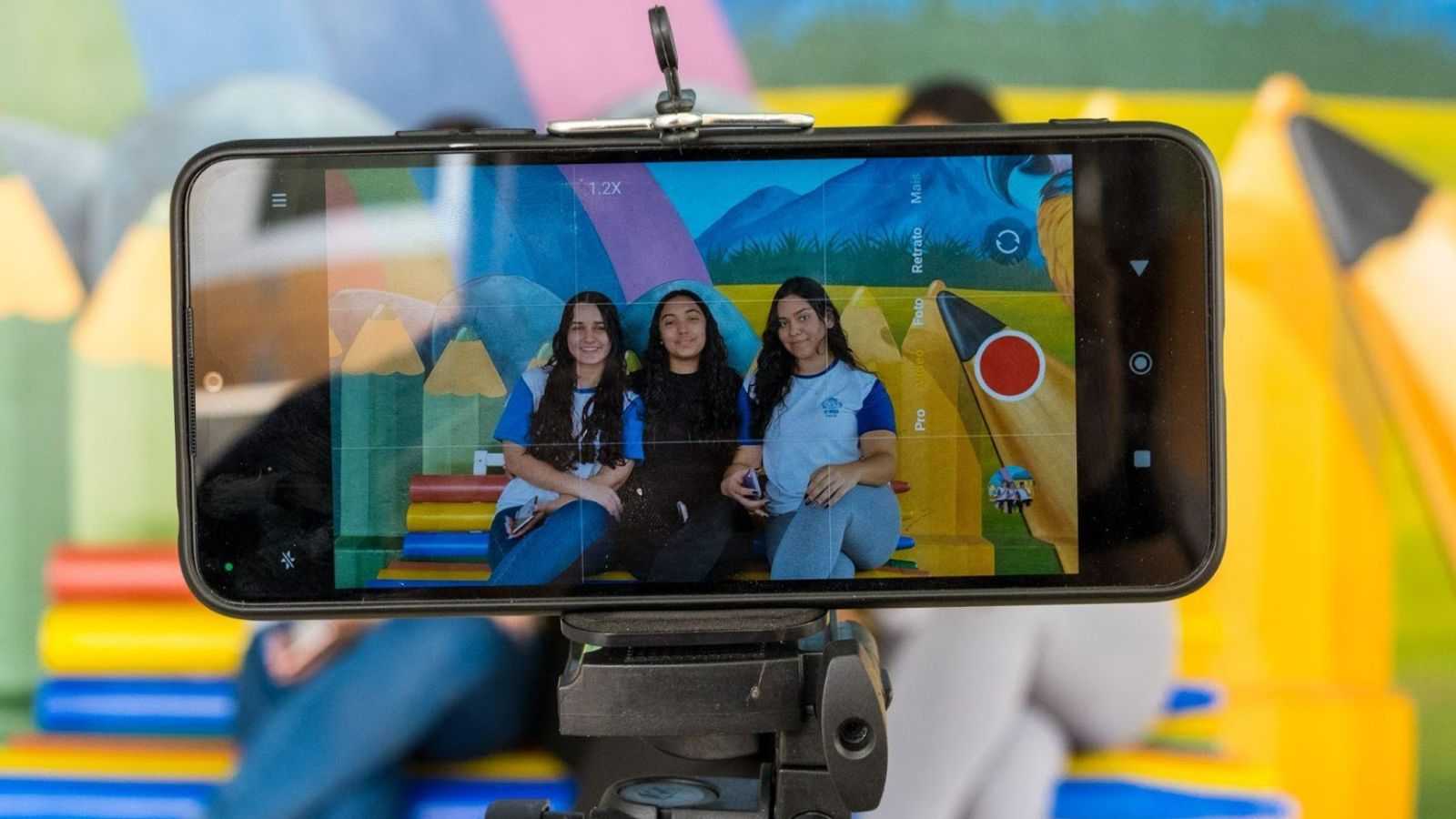 Celular em tripé grava três jovens estudantes sorrindo em frente a um mural colorido durante oficina de produção audiovisual em Jundiaí.