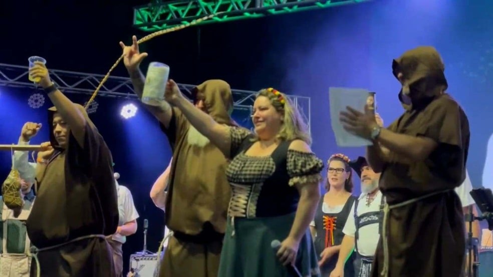 Participantes com trajes típicos alemães brindam no palco da Oktober Polo Jundiaí, segurando canecas de chope em clima de festa.