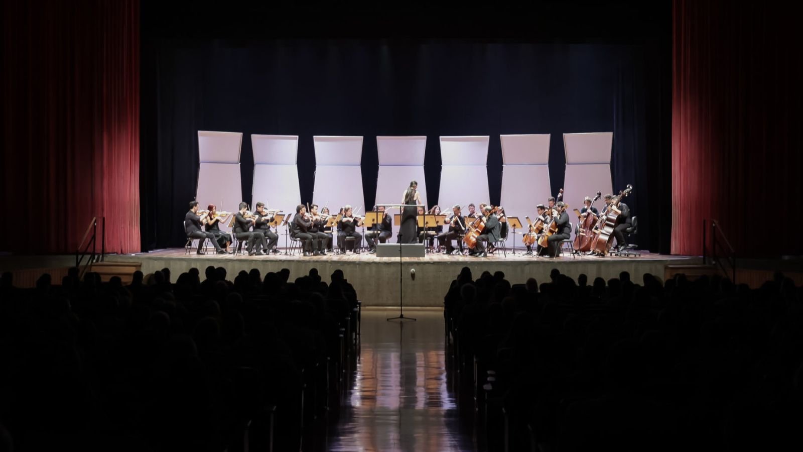 Orquestra Municipal de Jundiaí se apresenta no palco do Teatro Polytheama com regência da maestrina e músicos de cordas diante do público.