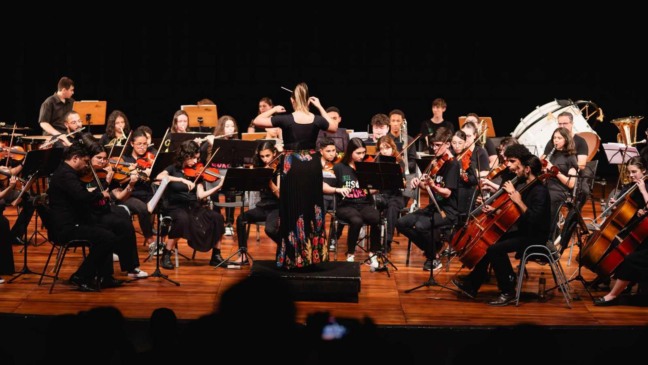 Orquestra Sinfônica do GURI Jundiaí em apresentação, com regente de costas e jovens músicos tocando instrumentos de cordas, sopro e percussão no palco.