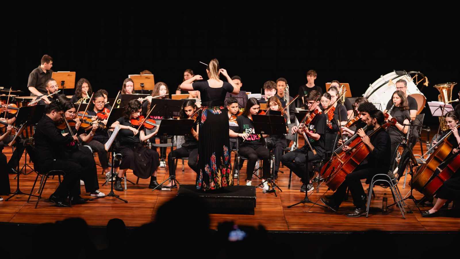 Orquestra Sinfônica do GURI Jundiaí em apresentação, com regente de costas e jovens músicos tocando instrumentos de cordas, sopro e percussão no palco.