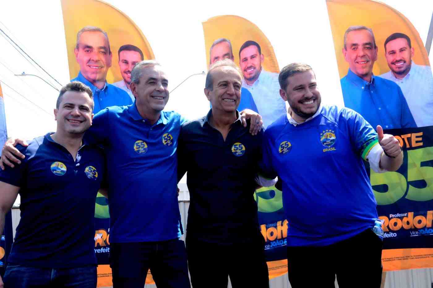Professor Rodolfo e equipe posam sorridentes em frente a banners de campanha durante evento em Várzea Paulista.