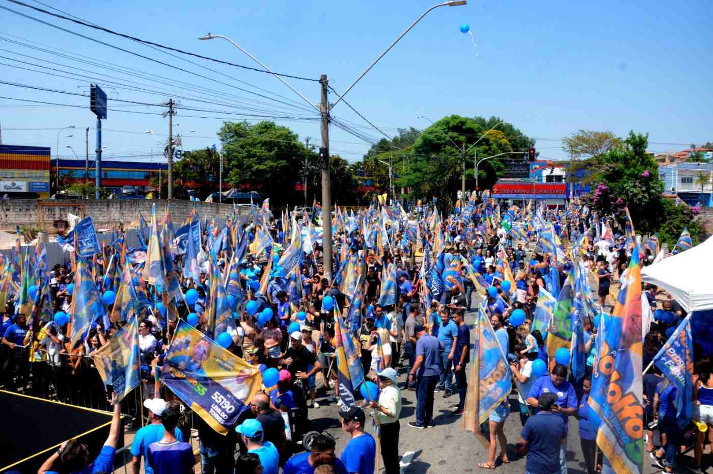 Multidão participa da Caminhada do Futuro com Professor Rodolfo em Várzea Paulista, segurando bandeiras e balões de campanha.