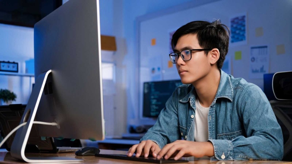 Programador usando óculos e jaqueta jeans digita no teclado enquanto trabalha em um computador em escritório de tecnologia. Programador usando óculos e jaqueta jeans digita no teclado enquanto trabalha em um computador em escritório de tecnologia.