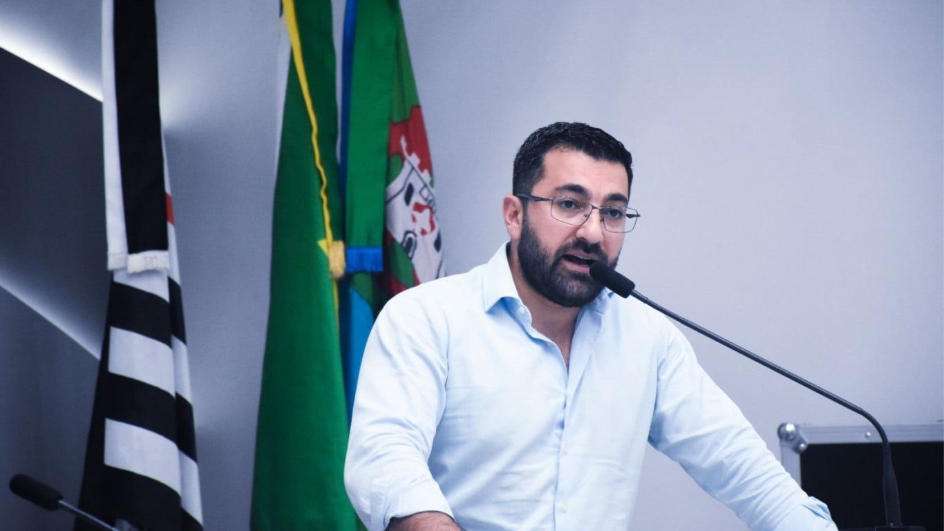 Vereador Faouaz Taha fala ao microfone na Câmara de Jundiaí, com bandeiras ao fundo.