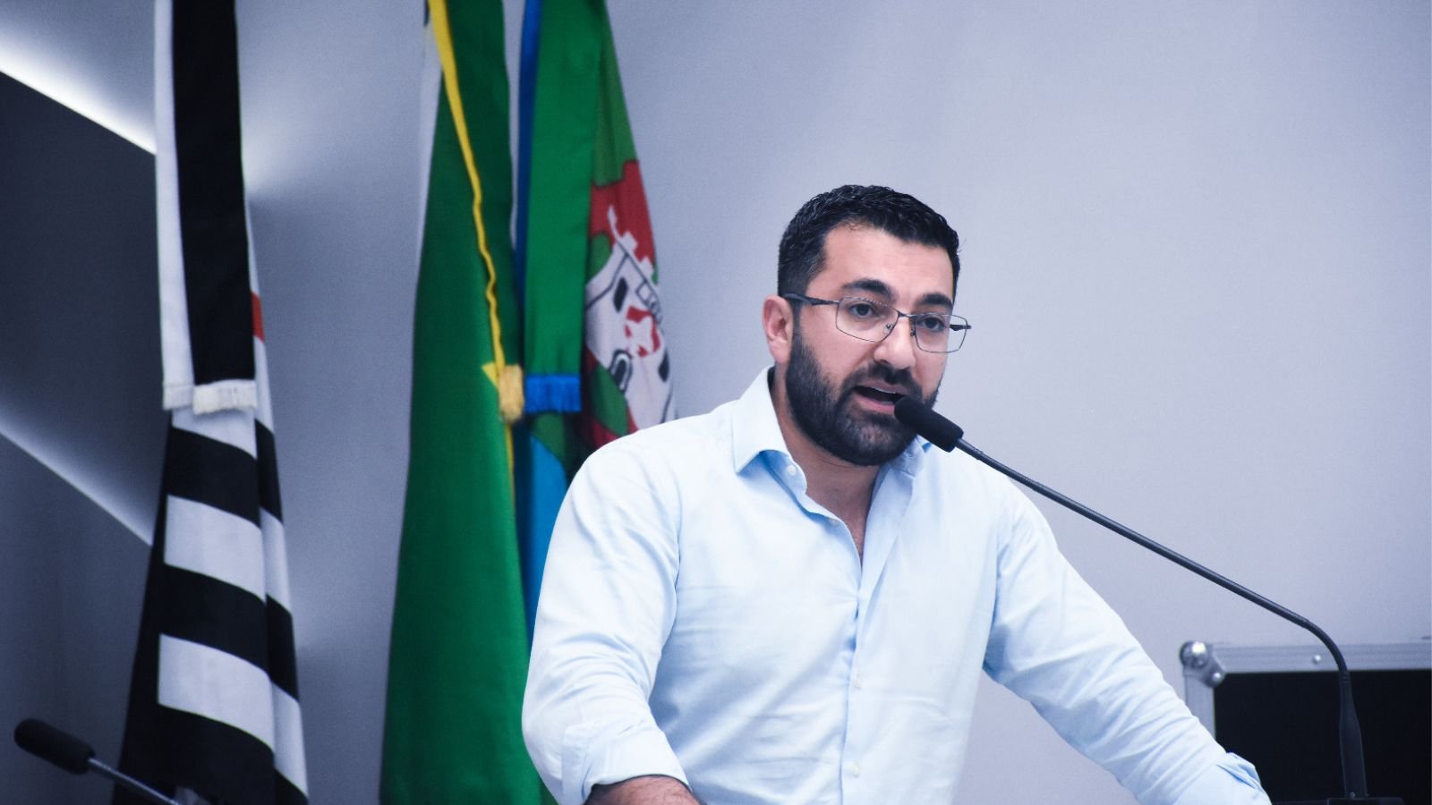 Vereador Faouaz Taha fala ao microfone na Câmara de Jundiaí, com bandeiras ao fundo.