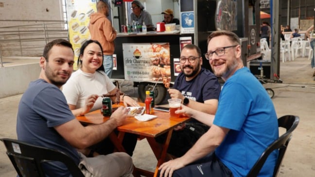 Grupo de quatro pessoas sentadas à mesa, sorrindo enquanto saboreiam espetinhos e bebidas artesanais no evento 5º Sabores da Gente, realizado no Espaço Expressa, em Jundiaí. Ao fundo, é possível ver um food truck especializado em churrasco.