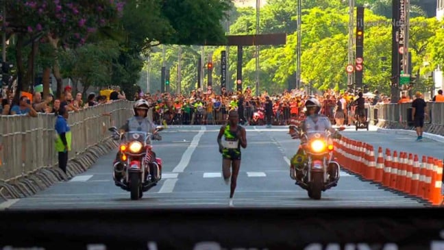 Corredora africana na liderança da São Silvestre 2024, cercado por motocicletas da polícia, público e vegetação ao fundo.