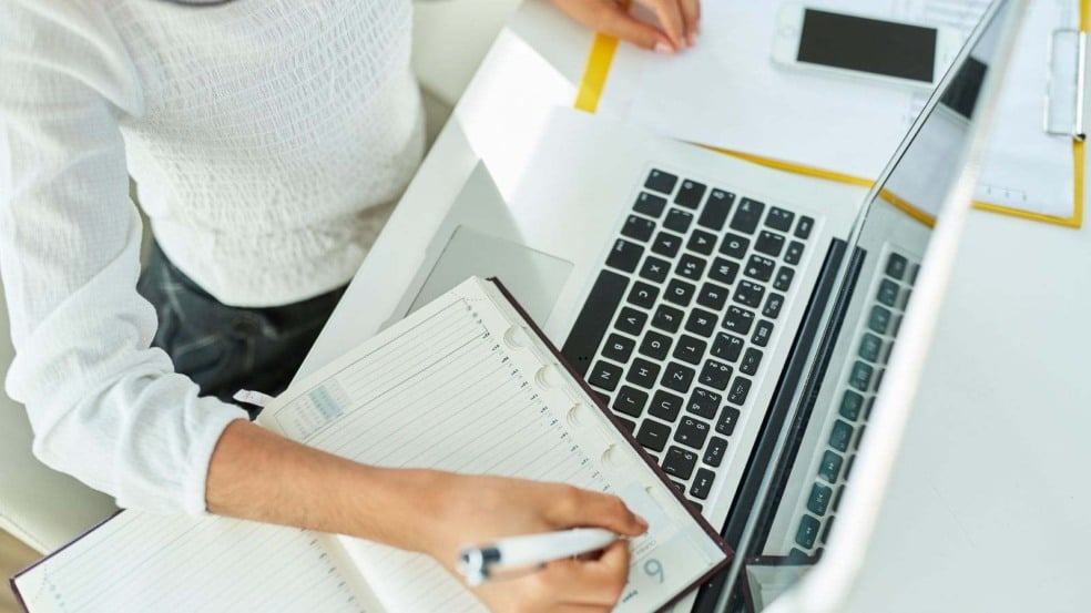 Profissional anota compromissos em um caderno ao lado de um notebook aberto, simbolizando planejamento e gestão de tarefas.