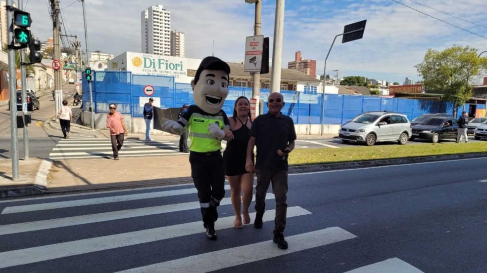 Mascote Vaguinho atravessa a faixa de pedestres em Jundiaí com duas pessoas, durante ação da Semana Nacional de Trânsito, enquanto carros aguardam no semáforo. Mascote Vaguinho atravessa a faixa de pedestres em Jundiaí com duas pessoas, durante ação da Semana Nacional de Trânsito, enquanto carros aguardam no semáforo.