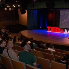 Vista geral da plateia assistindo à abertura do TEDxJundiaí, com destaque para o palco iluminado e o telão exibindo um palestrante. A ambientação mostra a atmosfera acolhedora do Centro das Artes de Jundiaí.