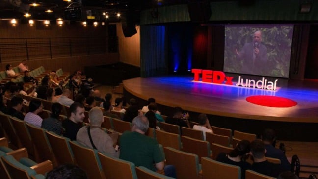 Vista geral da plateia assistindo à abertura do TEDxJundiaí, com destaque para o palco iluminado e o telão exibindo um palestrante. A ambientação mostra a atmosfera acolhedora do Centro das Artes de Jundiaí.