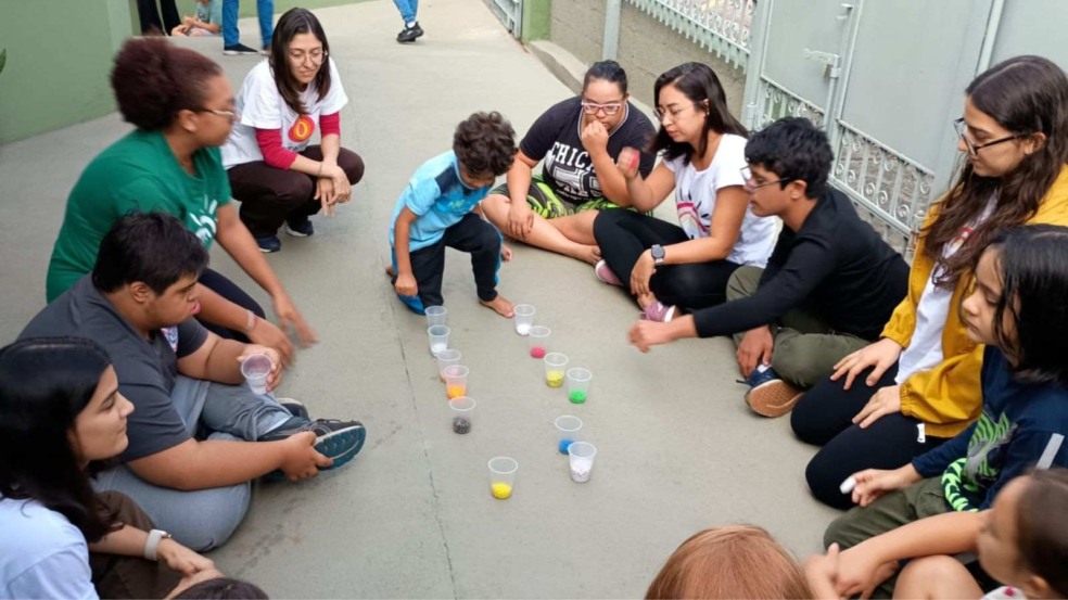 Pessoas reunidas em roda no chão participando de atividade lúdica com copos de cores variadas na Associação Teia Down em Jundiaí.