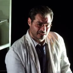 Thiago Lacerda em cena do espetáculo “A Peste”, usando jaleco branco e óculos, iluminado em palco escuro.