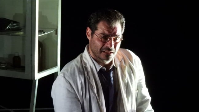 Thiago Lacerda em cena do espetáculo “A Peste”, usando jaleco branco e óculos, iluminado em palco escuro. Thiago Lacerda em cena do espetáculo “A Peste”, usando jaleco branco e óculos, iluminado em palco escuro.