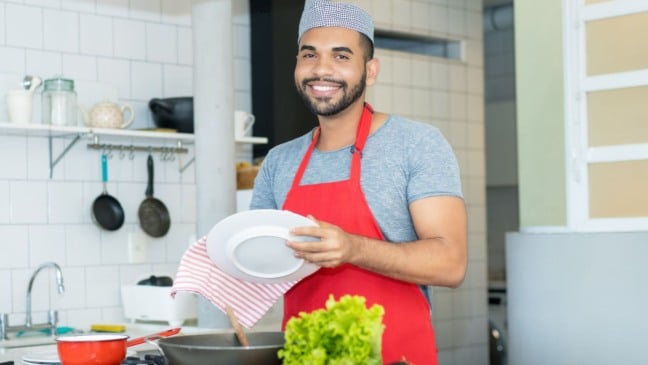 Homem sorridente com avental vermelho secando prato em cozinha industrial.