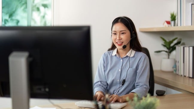 Mulher atendente de call center trabalhando no computador e sorrindo durante atendimento remoto em home office.
