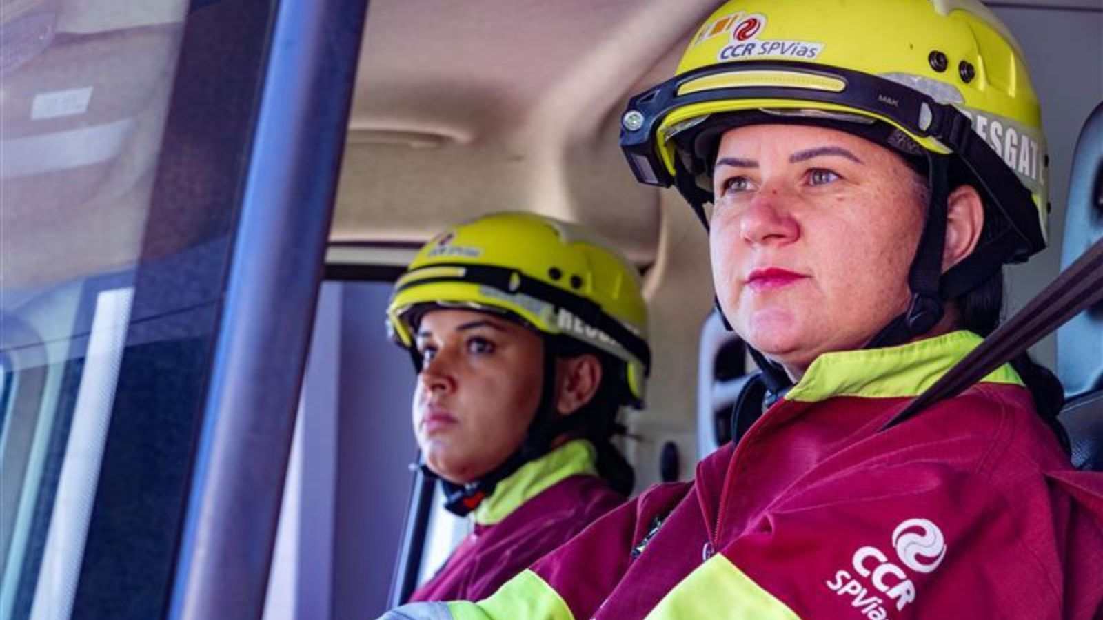Duas colaboradoras da Motiva, com uniformes e capacetes de resgate amarelos e vermelhos, dentro de um veículo operacional da AutoBAn, prontas para atendimento nas rodovias Anhanguera-Bandeirantes.