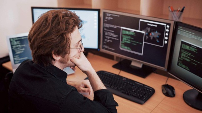 Programador analisa códigos em três monitores durante o trabalho em home office.