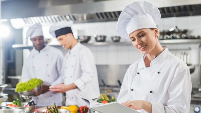 Cozinheiros uniformizados em cozinha industrial, preparando alimentos e consultando receitas.