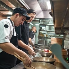 Cozinheiros preparando alimentos em uma cozinha profissional com ambiente industrial e utensílios de restaurante.