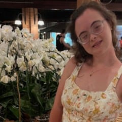 Jovem mulher com óculos e vestido floral posa sorridente ao lado de orquídeas brancas em ambiente interno bem iluminado.